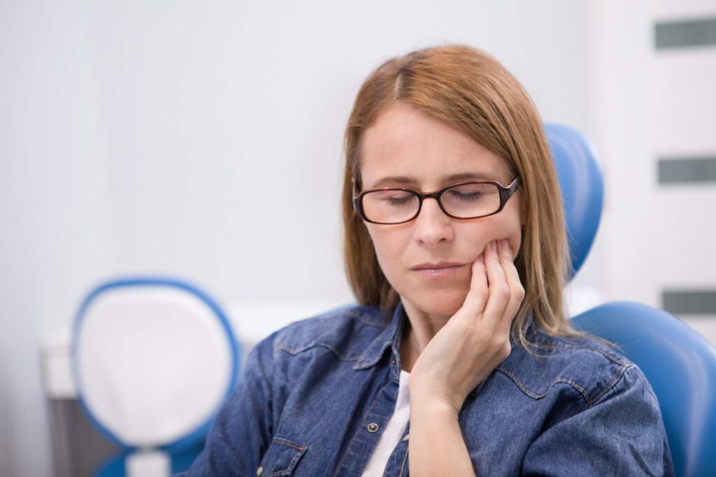 Mujer con gafas y la mano en la mandíbula sentada en el sillón del dentista